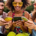 Feliz jovem companhia de amigos sorridentes sentados no parque usando smartphones homens e mulheres se divertindo juntos