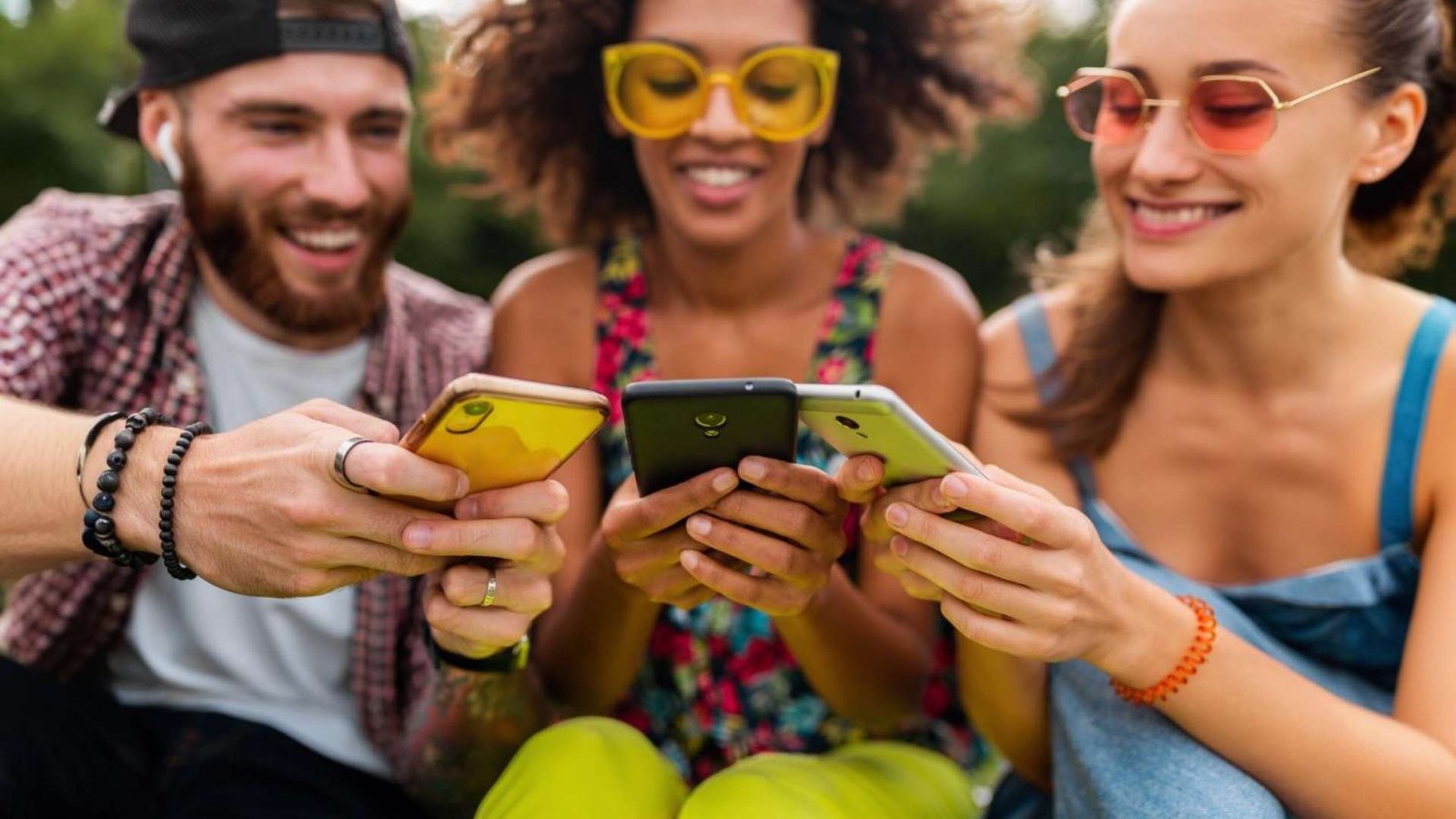 Feliz jovem companhia de amigos sorridentes sentados no parque usando smartphones homens e mulheres se divertindo juntos