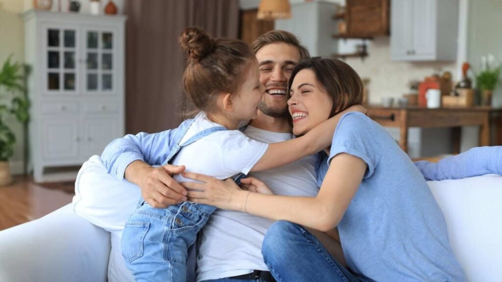 Pais jovens amigáveis positivos com a filha sorridente sentados no sofá juntos enquanto relaxam em casa no fim de semana
